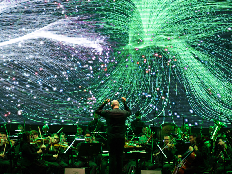 Concierto de la Orquesta Filarmónica de Medellín con una proyección visual de luces verdes y patrones abstractos en el fondo, creando una experiencia inmersiva entre música y tecnología.