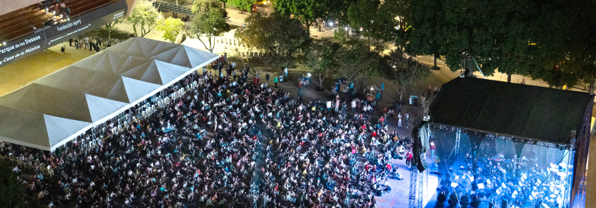 Vista aérea de un concierto al aire libre de la Orquesta Filarmónica de Medellín en el Parque de los Deseos, con una gran audiencia reunida en sillas y en el suelo, disfrutando de la presentación nocturna bajo una iluminación vibrante.