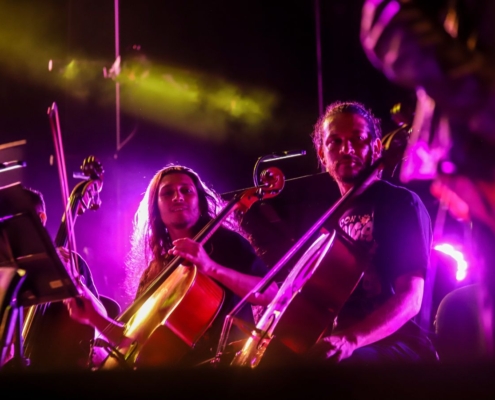 Músicos tocando violonchelo en un ambiente iluminado con luces púrpuras y amarillas, representando el talento de la Orquesta Filarmónica de Medellín.