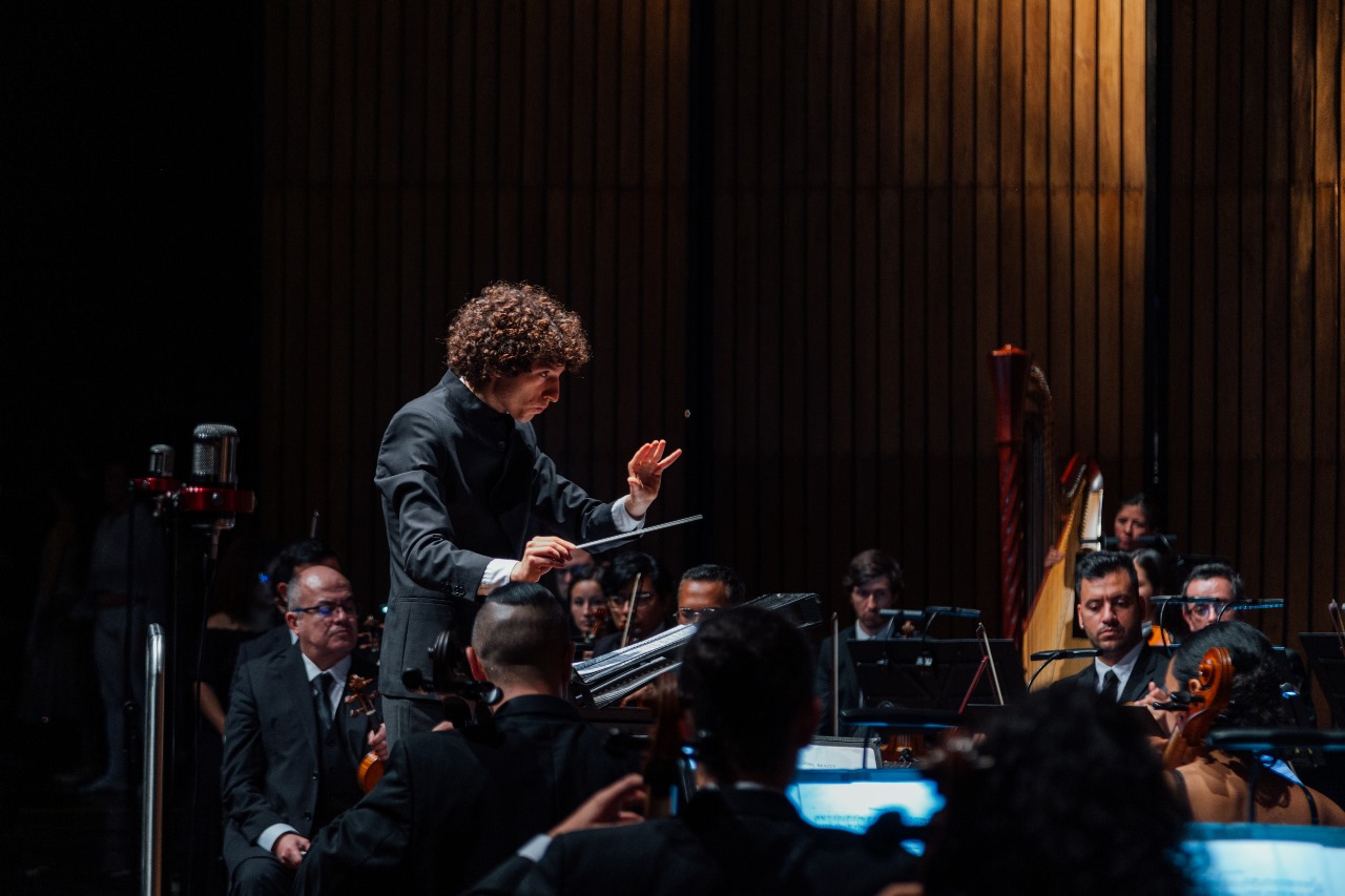 Tami Daniel Rueda Blanco dirigiendo la orquesta Filarmed durante un concierto en sala con acústica de madera.