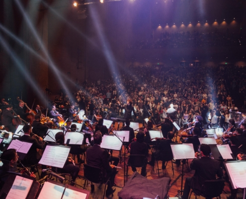 Vista desde el escenario de un concierto de la Orquesta Filarmónica de Medellín, con músicos tocando frente a un auditorio lleno de público de pie, envuelto en luces cálidas y ambiente festivo.