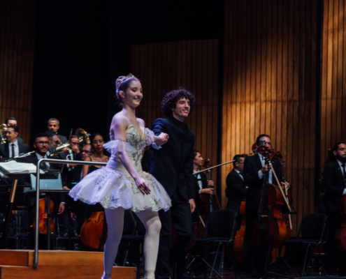 Bailarina y director musical saludan sonrientes al público tras una presentación sinfónica con la Orquesta Filarmónica de Medellín.