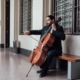 Músico de la Orquesta Filarmónica de Medellín interpretando el violonchelo en un pasillo institucional, sentado en una banca de madera.