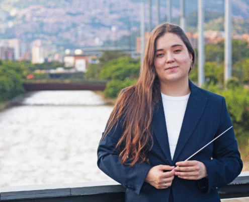 Ana María Patiño-Osorio sonríe mientras sostiene una batuta, con el río Medellín y la ciudad al fondo, en su presentación como directora titular de la Orquesta Filarmónica de Medellín.