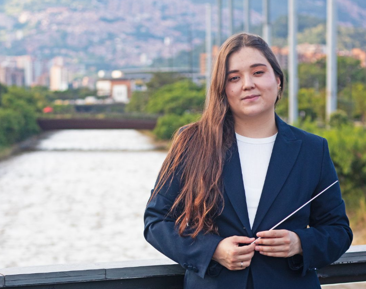 Ana María Patiño-Osorio sonríe mientras sostiene una batuta, con el río Medellín y la ciudad al fondo, en su presentación como directora titular de la Orquesta Filarmónica de Medellín.