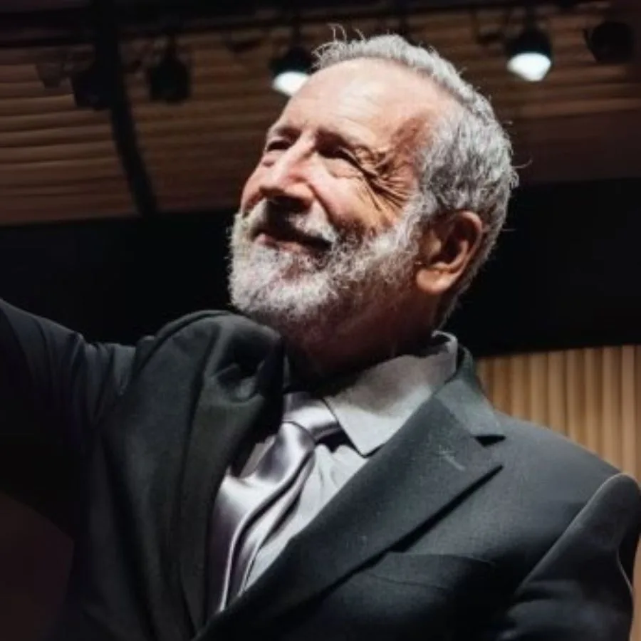 Director de orquesta veterano saludando al público en un auditorio, vestido con traje formal y corbata plateada.