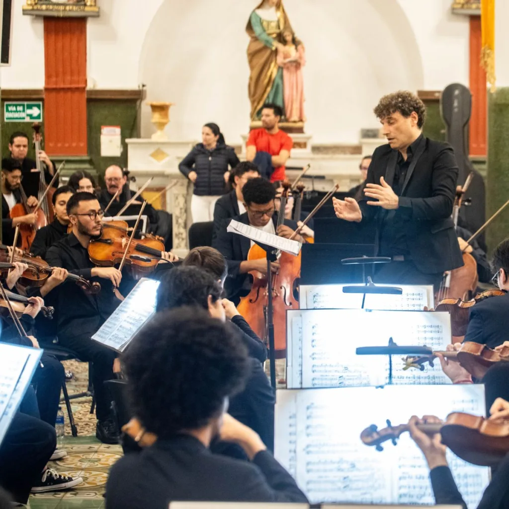 Director de orquesta lidera concierto sinfónico en iglesia con músicos jóvenes durante una presentación comunitaria en Antioquia.
