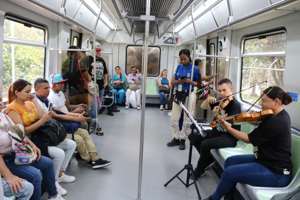 Músicos tocan violines en un vagón de metro mientras pasajeros los escuchan atentos, como parte de un programa de salud mental y bienestar.