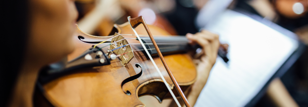 Mano de un músico sosteniendo un violín durante un ensayo orquestal, con músicos desenfocados al fondo