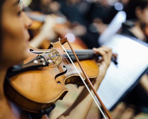 Mano de un músico sosteniendo un violín durante un ensayo orquestal, con músicos desenfocados al fondo