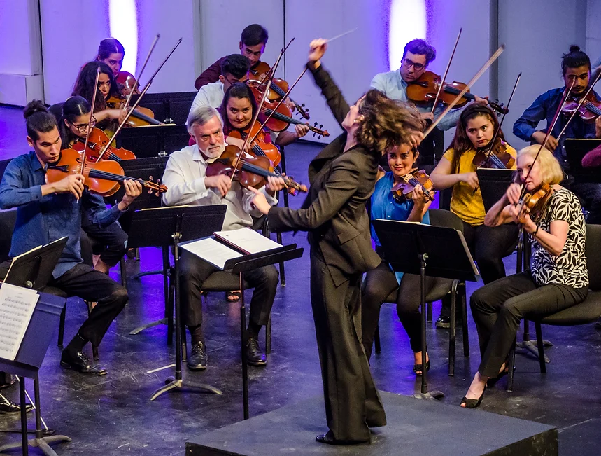 Alejandra Urrutia dirigiendo una orquesta de cuerdas en concierto, con músicos en plena ejecución