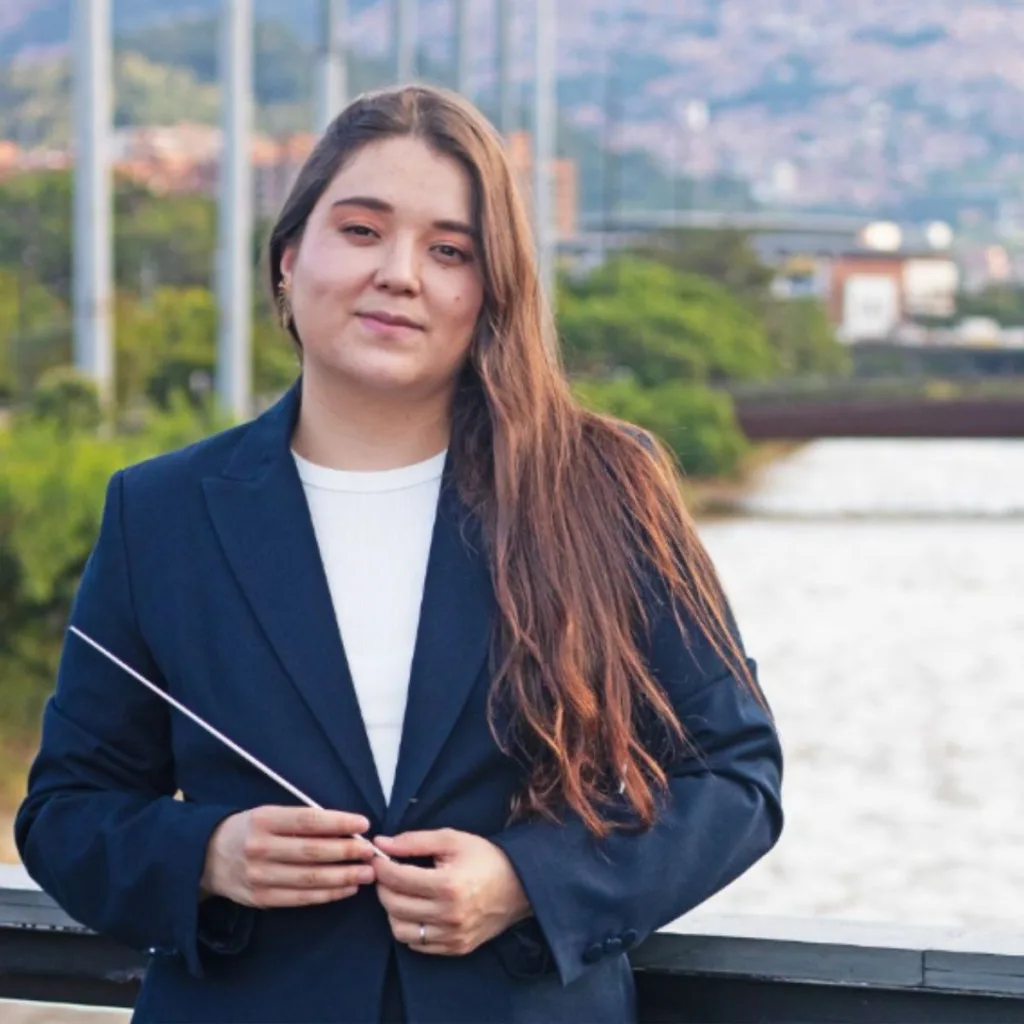 Ana María Patiño-Osorio sonríe mientras sostiene una batuta, con el río Medellín y la ciudad al fondo, en su presentación como directora titular de la Orquesta Filarmónica de Medellín.