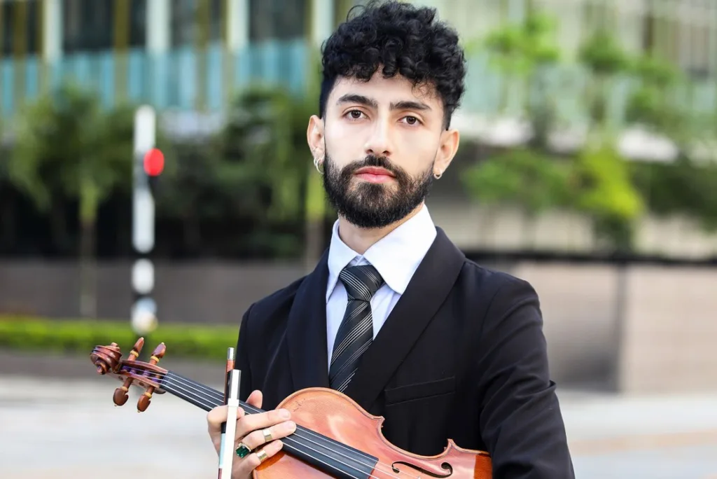 Camilo Martínez, joven violinista de la Filarmónica de Medellín, posa con su violín en traje formal frente a un entorno urbano moderno.