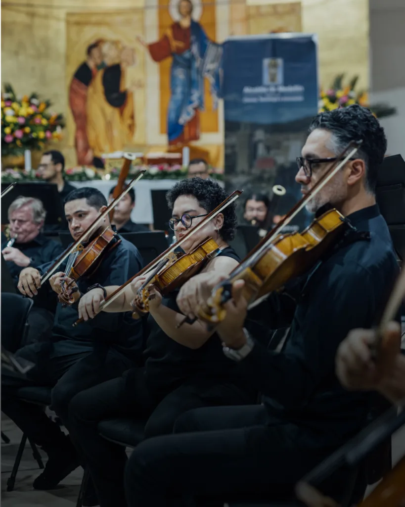 Músicos de cuerda de la Orquesta Filarmónica de Medellín interpretando en vivo durante un concierto en una iglesia, con un mural religioso de fondo.