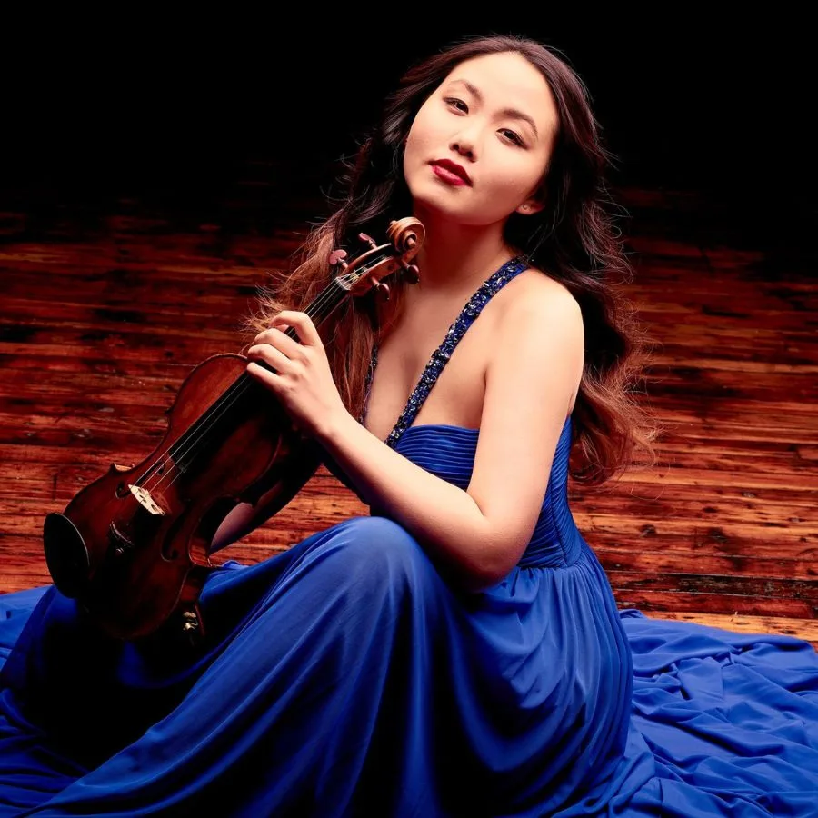 Retrato de Stella Chen sosteniendo su violín, con vestido azul y fondo de madera