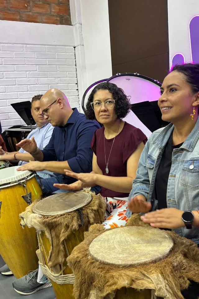 Personas de una empresa participan en un taller vivencial de liderazgo de la Orquesta Filarmónica de Medellín, tocando tambores en grupo.