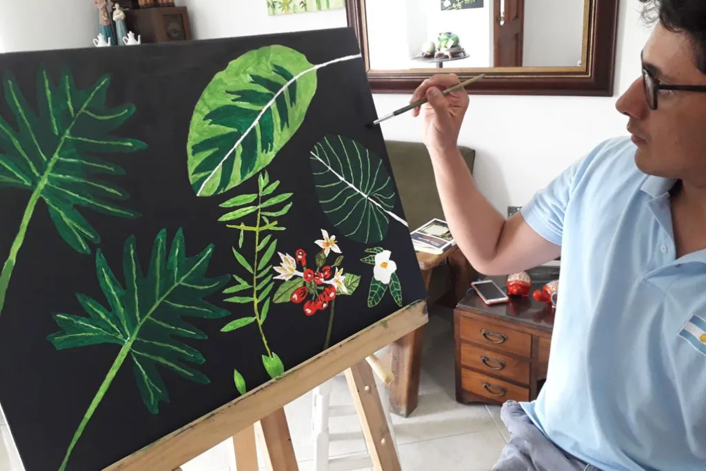 Tomás pintando una obra botánica sobre fondo negro, con hojas y flores tropicales