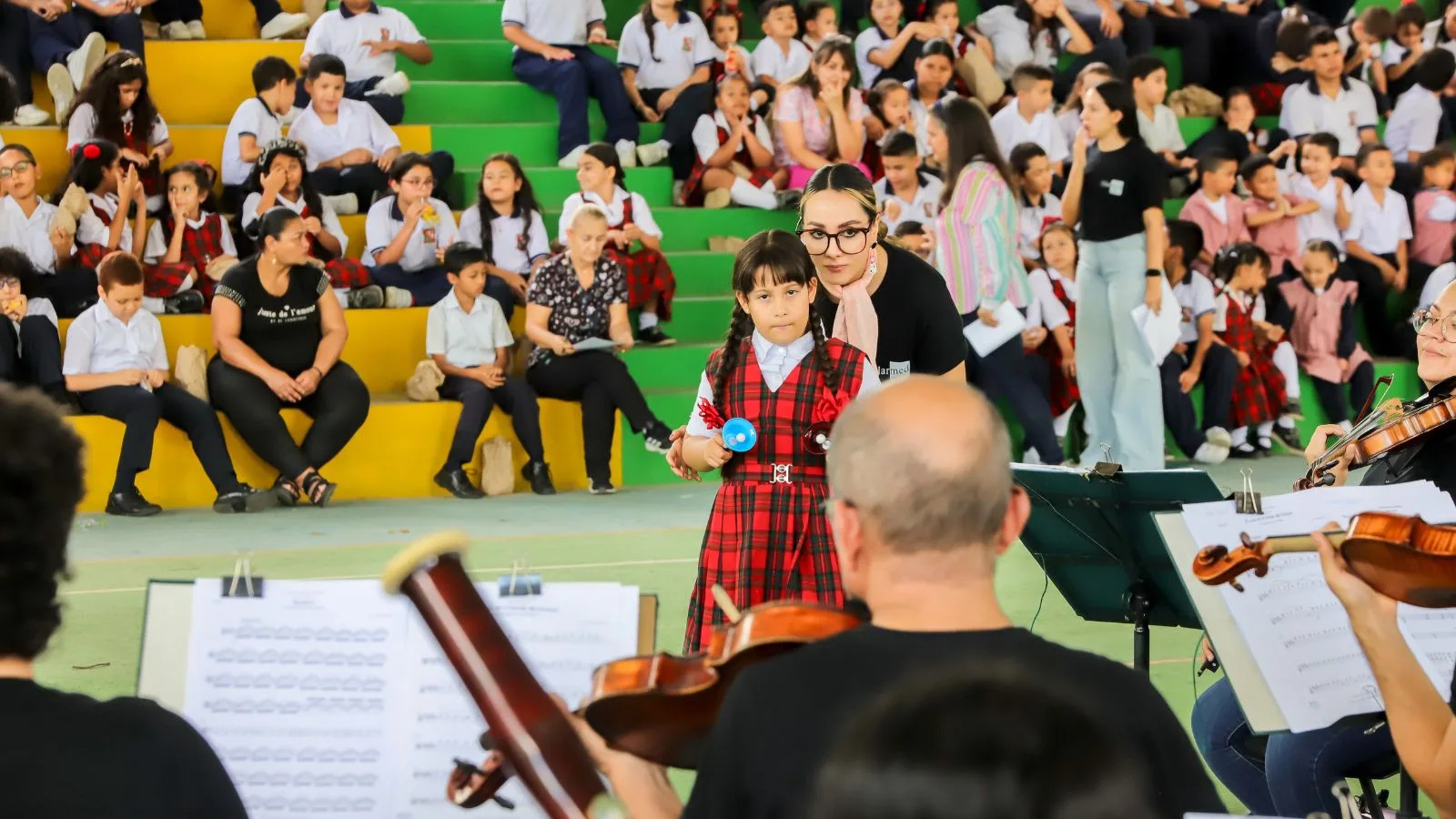 Niña con uniforme escolar participa tocando una maraca junto a músicos de la Orquesta Filarmónica de Medellín durante una actividad del programa FilU en su colegio.
