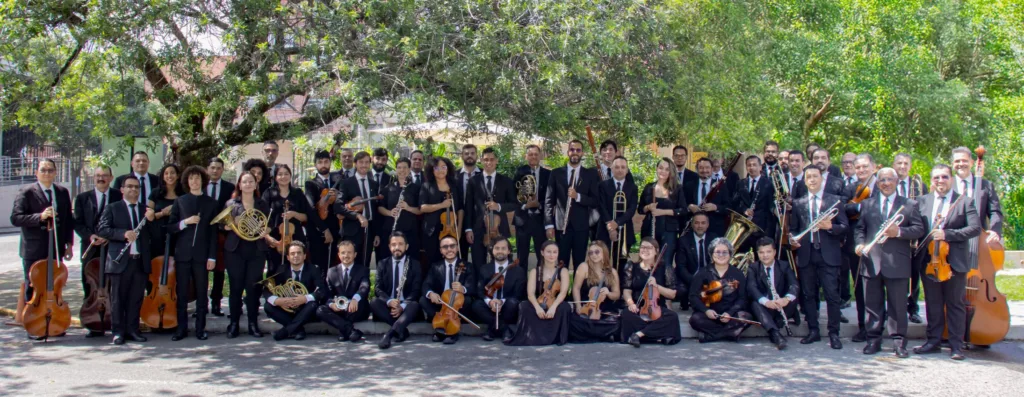 Todos los músicos que integran la Orquesta Filarmónica de Medellín posan sonriendo junto con su respectivo instrumento.