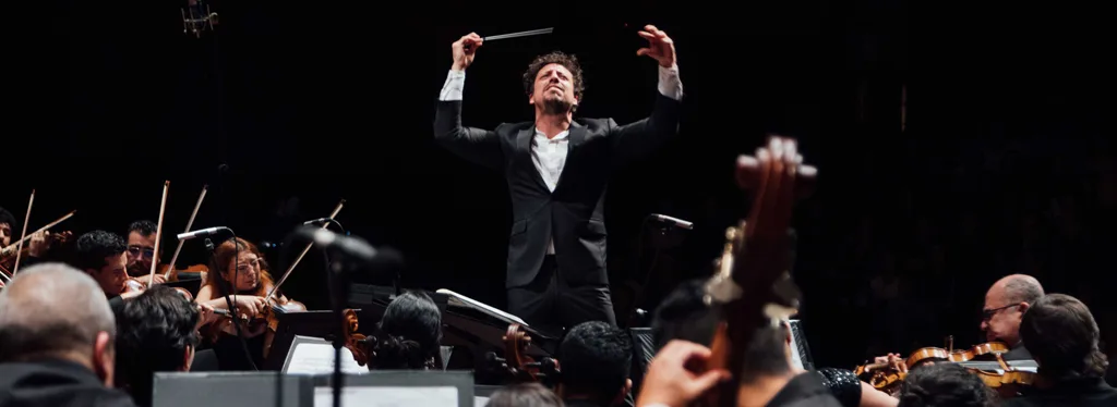 Paolo Bortolameoll concentrado dirigiendo una sinfonía, con la batuta levantada en un auditorio.