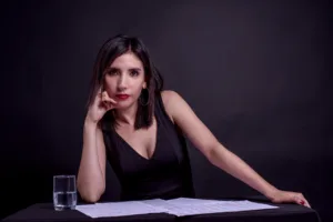 Mujer sentada frente a una mesa cubierta con un mantel negro. Sobre la mesa hay varias hojas con partituras musicales y un vaso de agua a su izquierda. La mujer tiene el cabello liso, corto y oscuro, lleva un vestido negro sin mangas y pendientes circulares. Apoya su cabeza ligeramente sobre su mano derecha mientras mira de frente a la cámara con una expresión seria y concentrada. El fondo es completamente oscuro, lo que dirige toda la atención hacia ella.