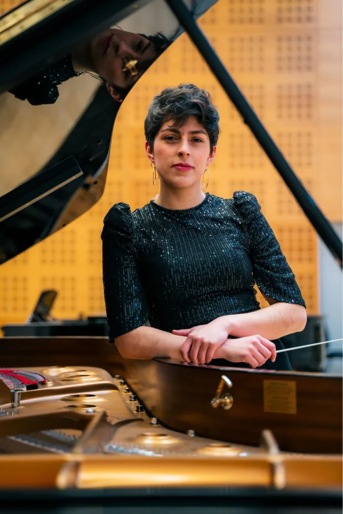 María Camila Barbosa, directora de orquesta, retratada junto a un piano de cola abierto, con vestuario oscuro y expresión concentrada, en un espacio de ensayo o concierto.